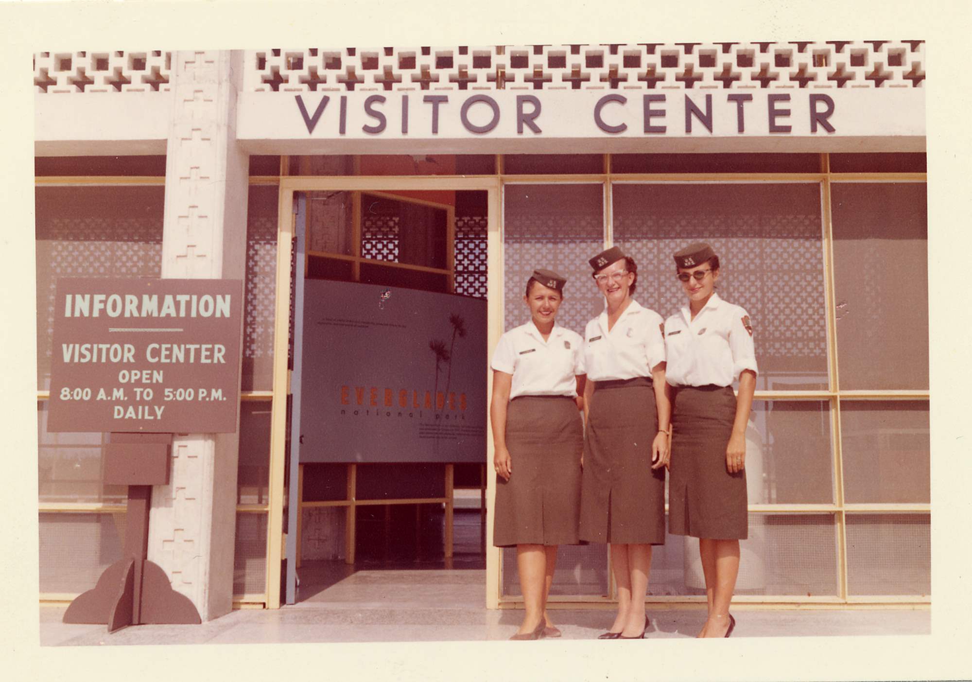 Three women stand in front of a visitor center building. They wear NPS uniform skirts, blouses, shield-shaped badges, and pillbox hats.